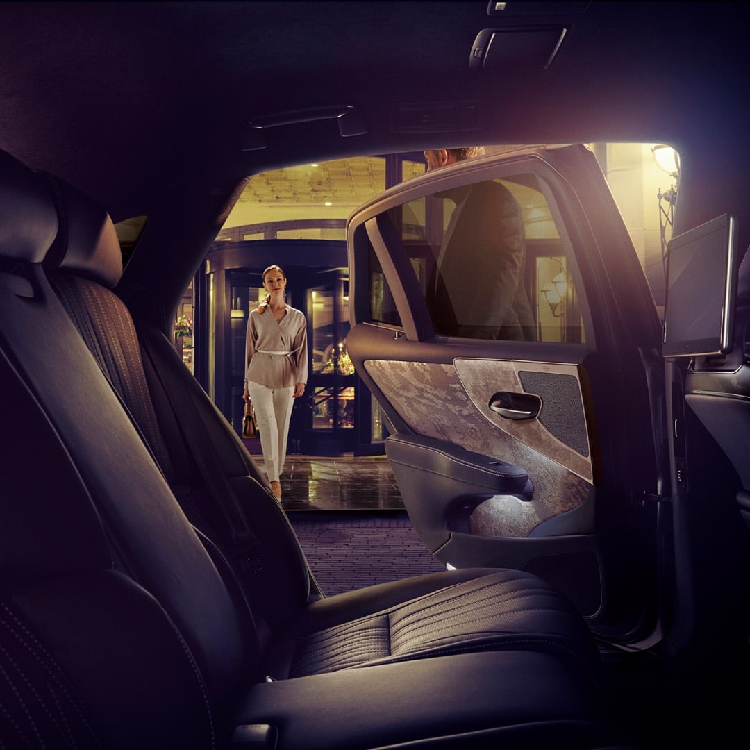 Wide shot from inside a Lexus LS, of a woman as she walks toward the LS. The back door is being held open by a man.